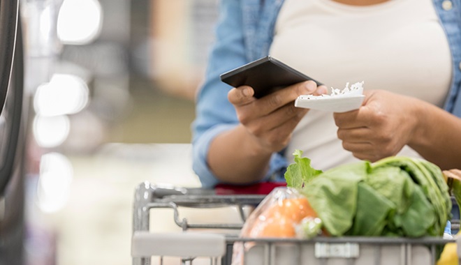 person with shopping trolley compariing shopping list with price on phone